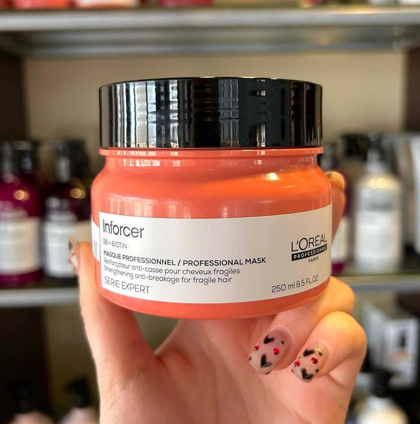 L'Oreal Serie Expert Inforcer hair mask held up in hand with shelves of hair products in the background.
